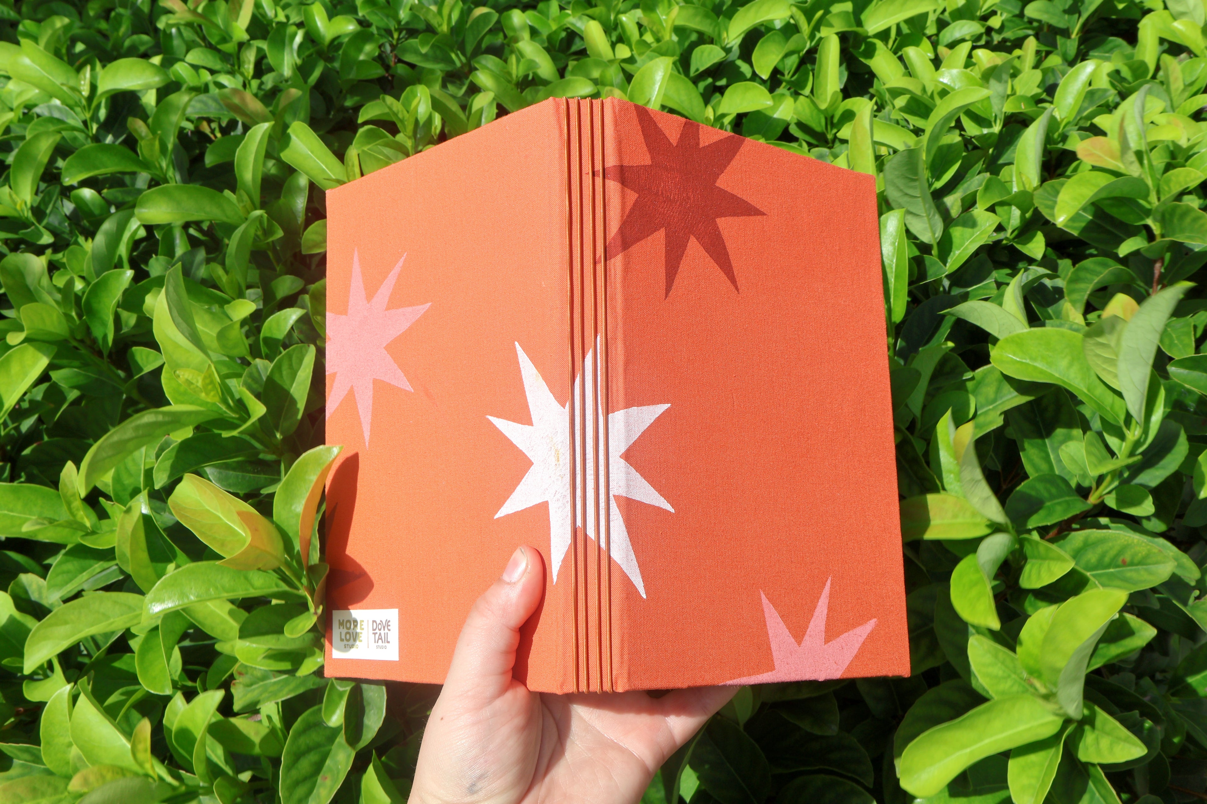A hand holds a folded orange hardcover refillable journal in front of dense green foliage. The cover has starburst patterns in white, pink, and dark red, with a small white label near the bottom left. hand bound and linocut printed with more love studio