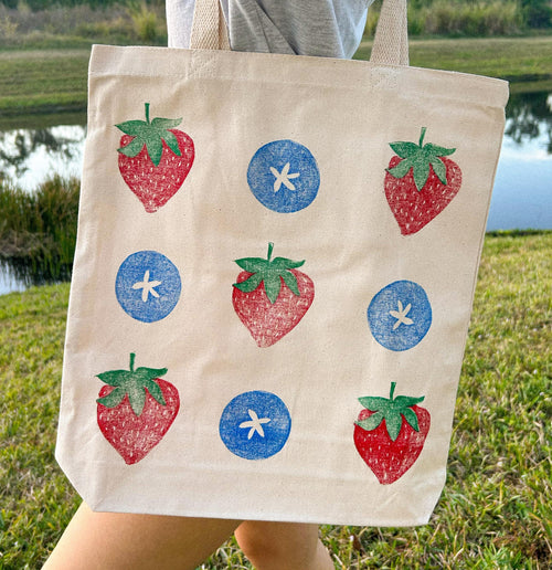 pattern showing red and green strawberries and blueberries printed on a cotton totebag. handmade linocut print green, red, and blue on canvas being worn by a person walking around a pond