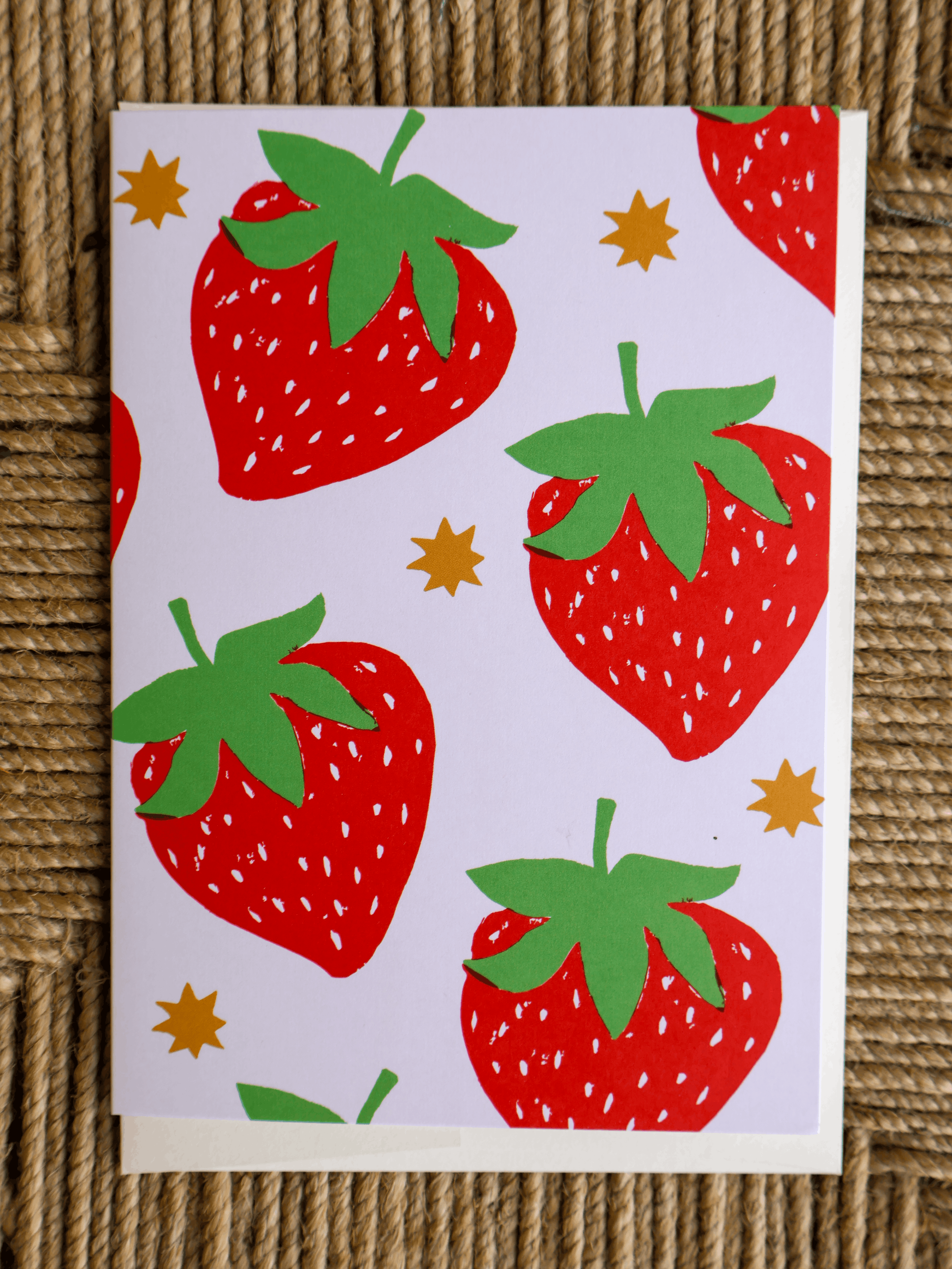 A square greeting card with a playful strawberry pattern, featuring red strawberries with green leaves and small gold stars on a white background, displayed on a textured brown woven mat.