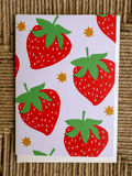 A square greeting card with a playful strawberry pattern, featuring red strawberries with green leaves and small gold stars on a white background, displayed on a textured brown woven mat.