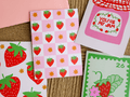 A styled flat-lay of strawberry-themed stationery, including a pink checkered card with red strawberries and gold stars, a white card reading 'YOU'RE MY JAM' in pink, a green postage-stamp-style card with a strawberry, and partial pink and white envelopes on a woven mat background.