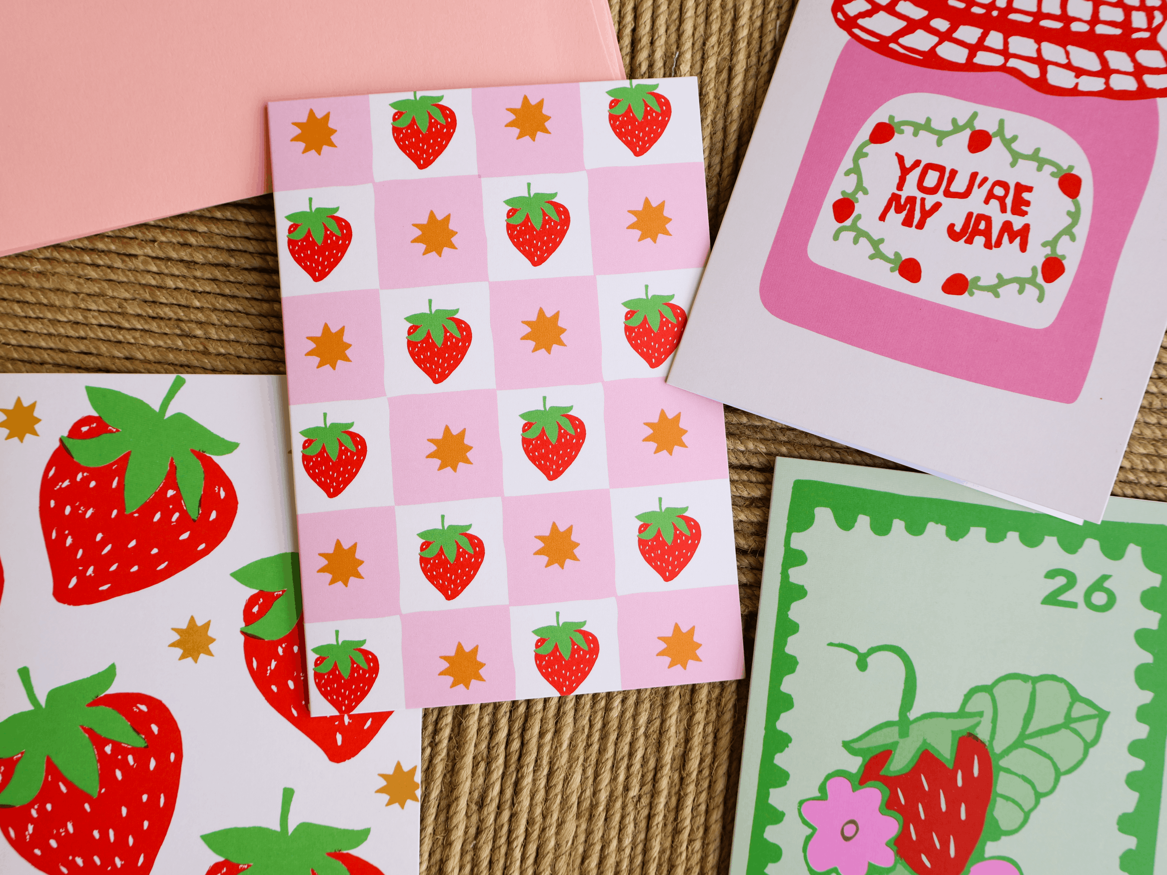 A styled flat-lay of strawberry-themed stationery, including a pink checkered card with red strawberries and gold stars, a white card reading 'YOU'RE MY JAM' in pink, a green postage-stamp-style card with a strawberry, and partial pink and white envelopes on a woven mat background.