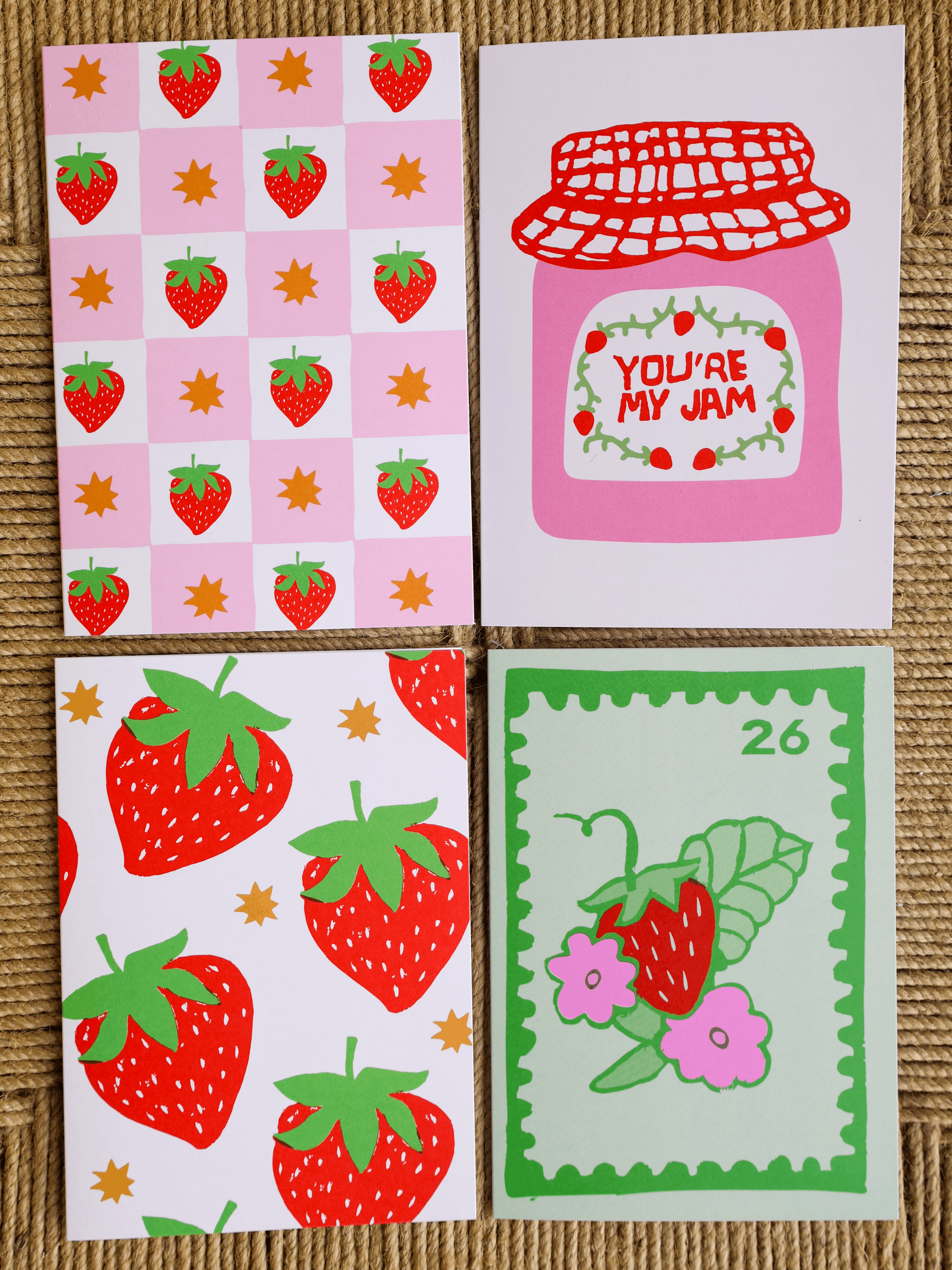 Four square greeting cards with strawberry-themed illustrations arranged in a 2x2 grid on a woven beige surface. The top-left card has a pink checkerboard pattern with red strawberries and small gold stars.