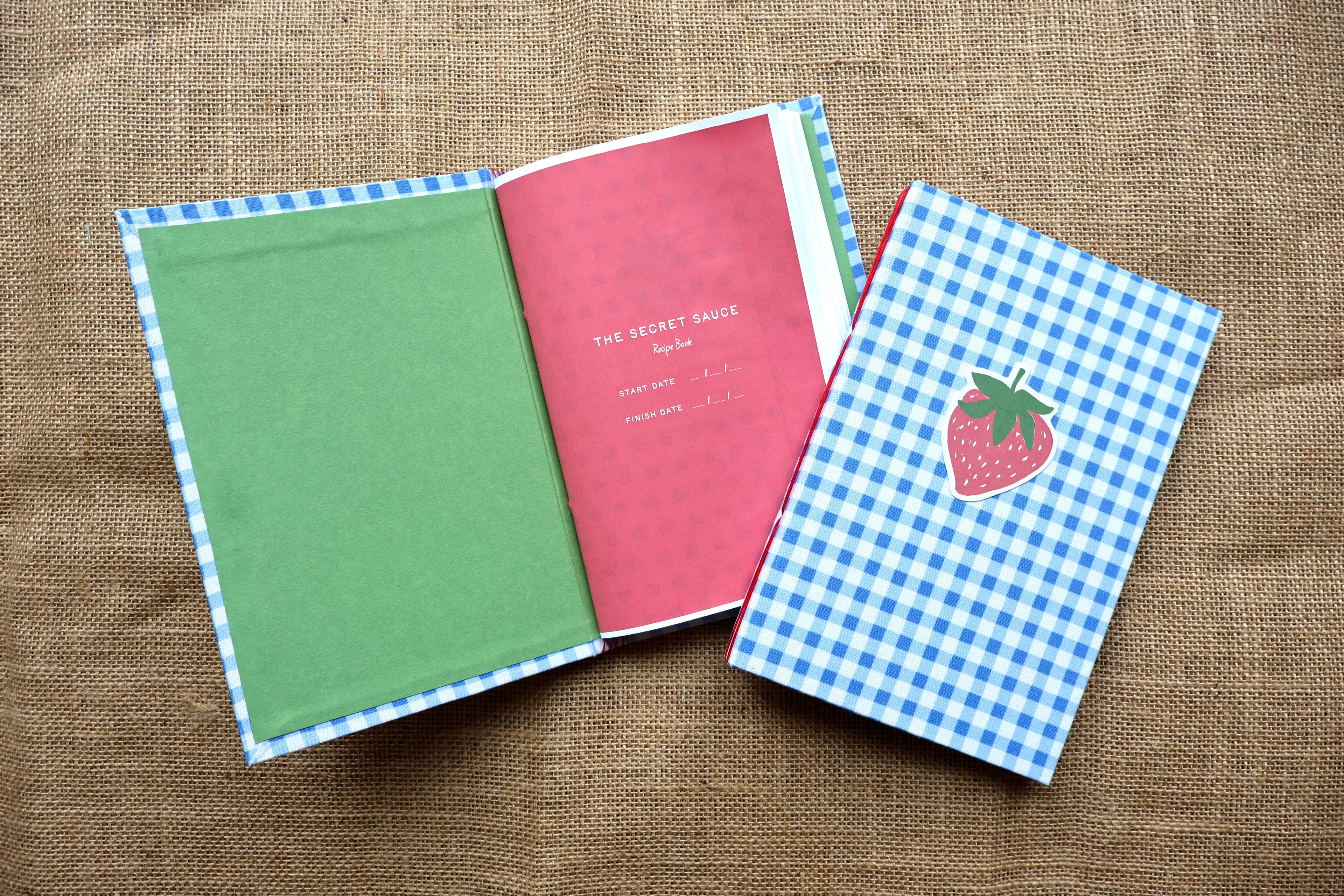 A handmade recipe book set displayed on a burlap background. One book is open, showing a green cover page and a pink interior page with the title "The Secret Sauce Recipe Book", while the other closed book features a blue-and-white gingham pattern cover and a strawberry illustration.