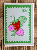 A square art print styled like a vintage postage stamp, featuring a red strawberry, pink flowers, and green leaves on a light gray background with a green border. The print is displayed flat on a textured brown woven mat.