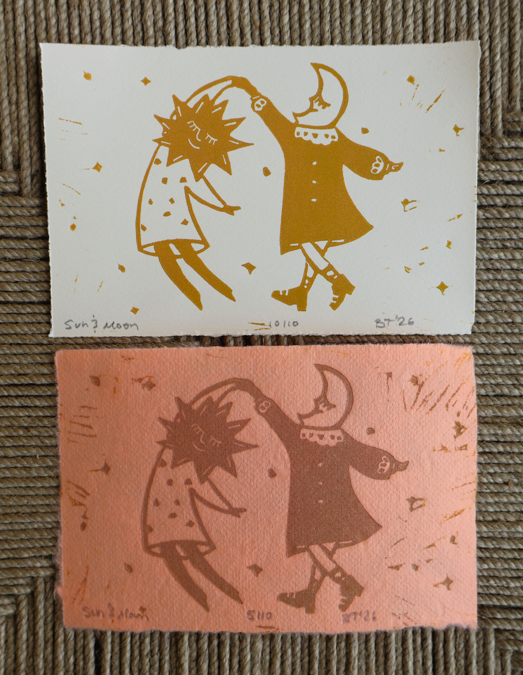 Two square art prints laid flat on a woven brown surface. The top print is white with gold linocut-style illustrations of a dancing sun and moon figure.