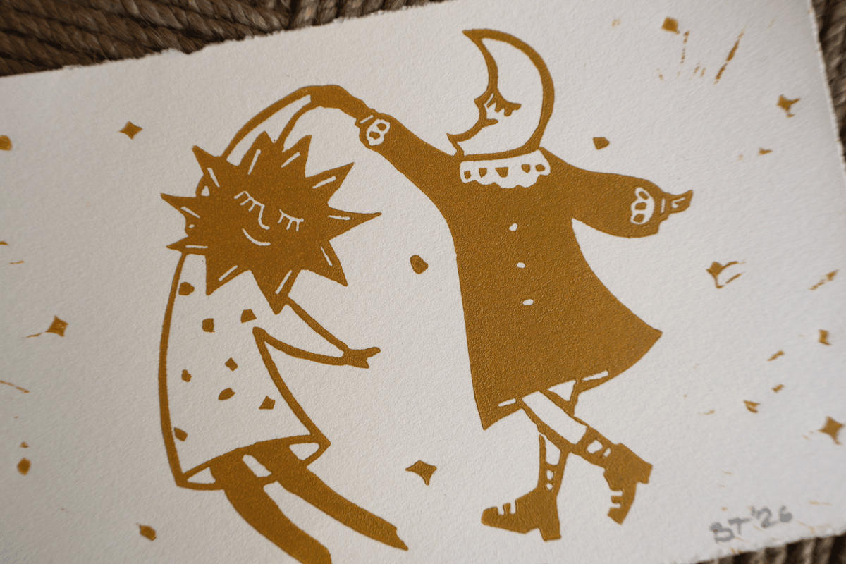 A close-up of a white art print featuring a stylized gold illustration of a dancing sun and moon figure, with small scattered gold marks on the paper.