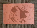 A square pink handmade paper art print featuring a brown linocut-style illustration of a sun and moon figure dancing, placed on a woven gray-green textured mat.