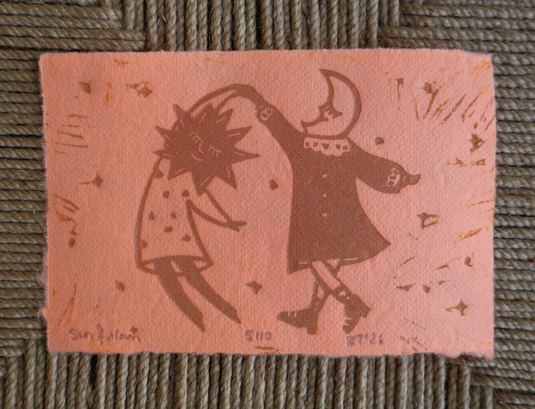 A square pink handmade paper art print featuring a brown linocut-style illustration of a sun and moon figure dancing, placed on a woven gray-green textured mat.