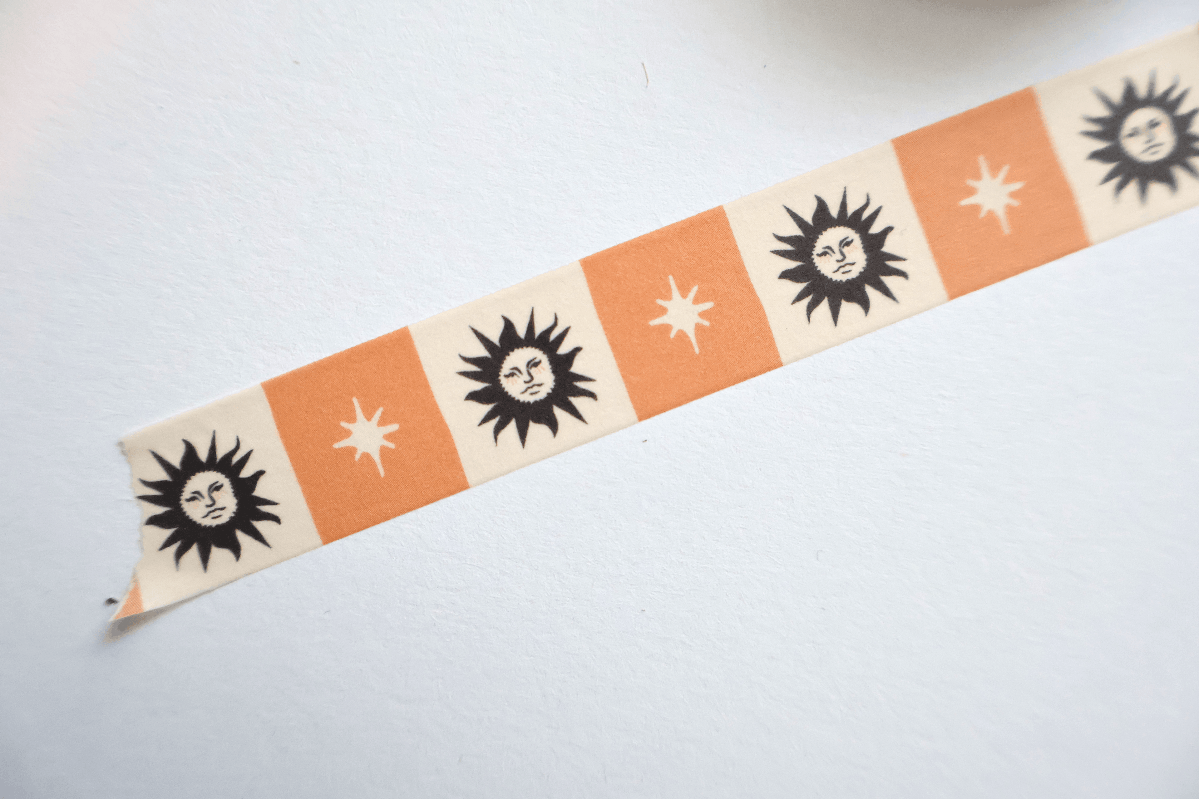A close-up of a decorative washi tape strip laid diagonally on a white surface. The tape features alternating orange and cream squares with black sun and starburst motifs.