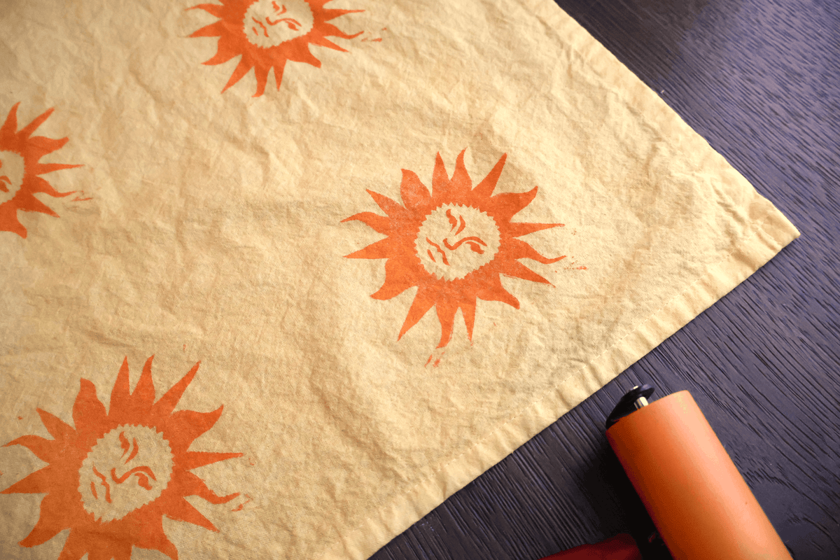 A close-up of a beige fabric item, likely a napkin or placemat, printed with orange sun motifs, laid diagonally on a dark wood surface with an orange pen visible in the corner.