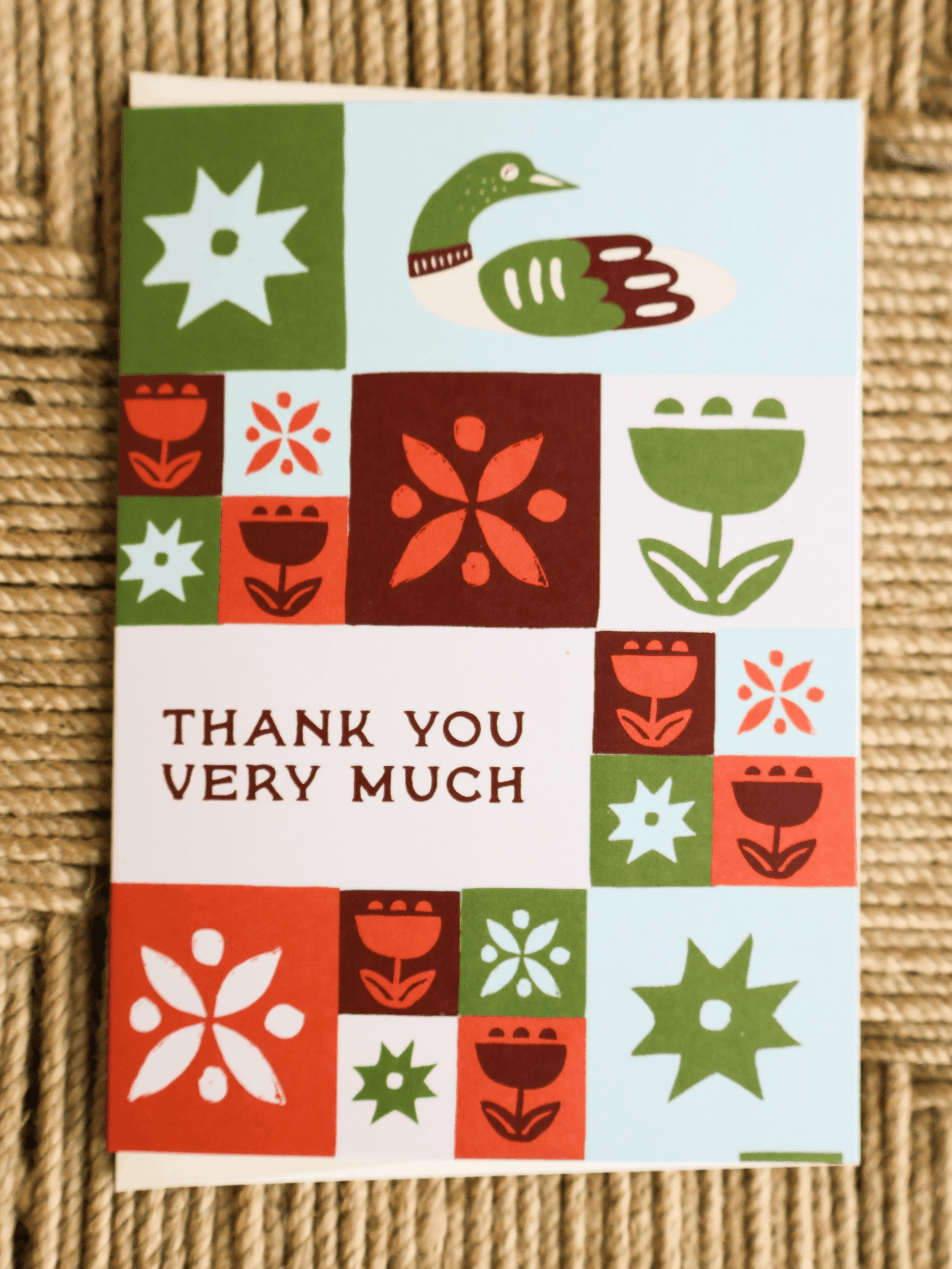 A square greeting card with a retro patchwork design in green, red, orange, and light blue, featuring a duck and floral motifs, with the text 'THANK YOU VERY MUCH' printed on the front. The card is photographed on a textured beige woven surface.