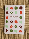A square greeting card with a white background, featuring a grid of colorful starburst shapes in red, green, and brown, with the text 'THANK YOU SO MUCH!' in the center. The card is photographed flat on a woven beige surface.