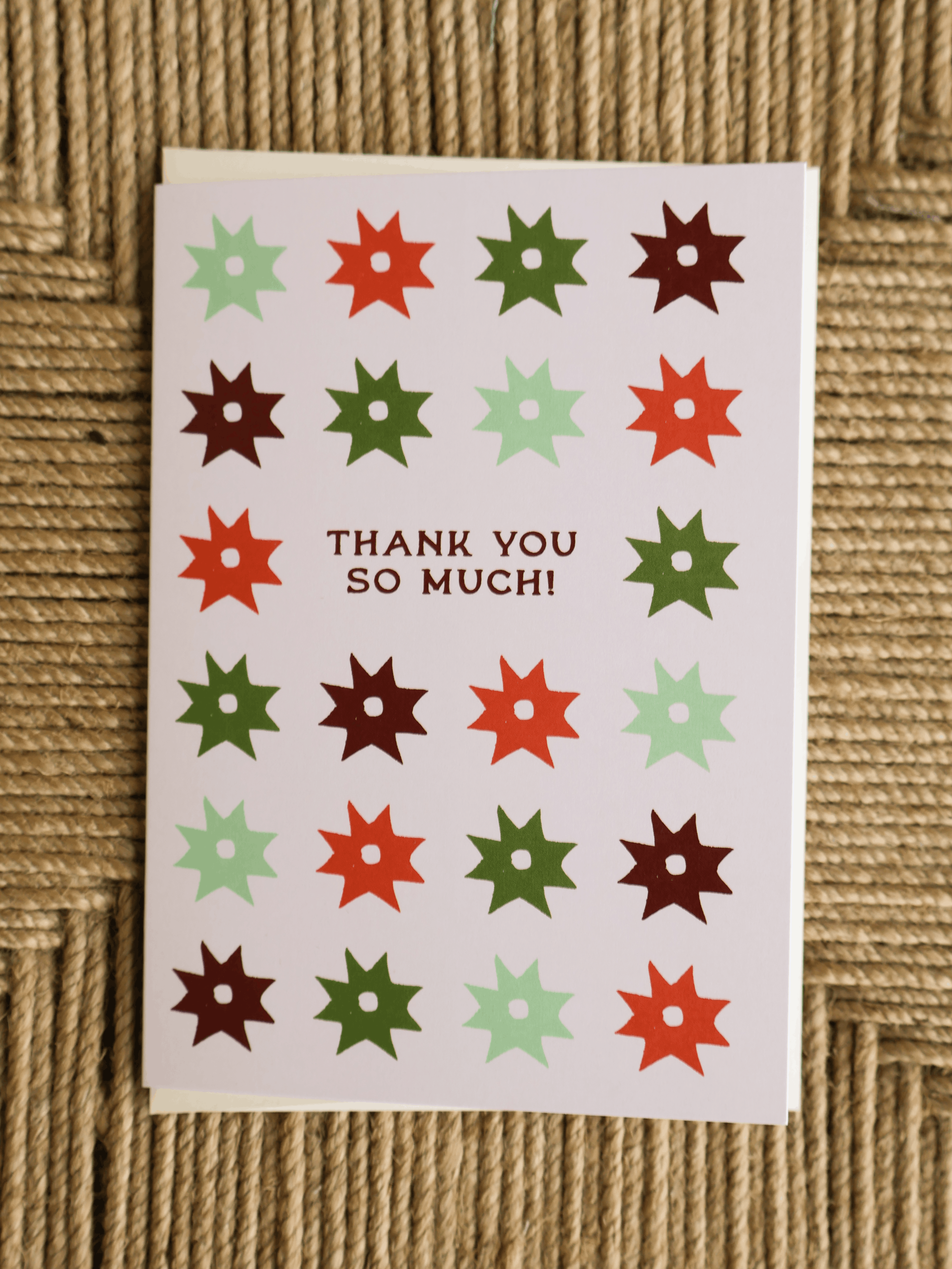 A square greeting card with a white background, featuring a grid of colorful starburst shapes in red, green, and brown, with the text 'THANK YOU SO MUCH!' in the center. The card is photographed flat on a woven beige surface.
