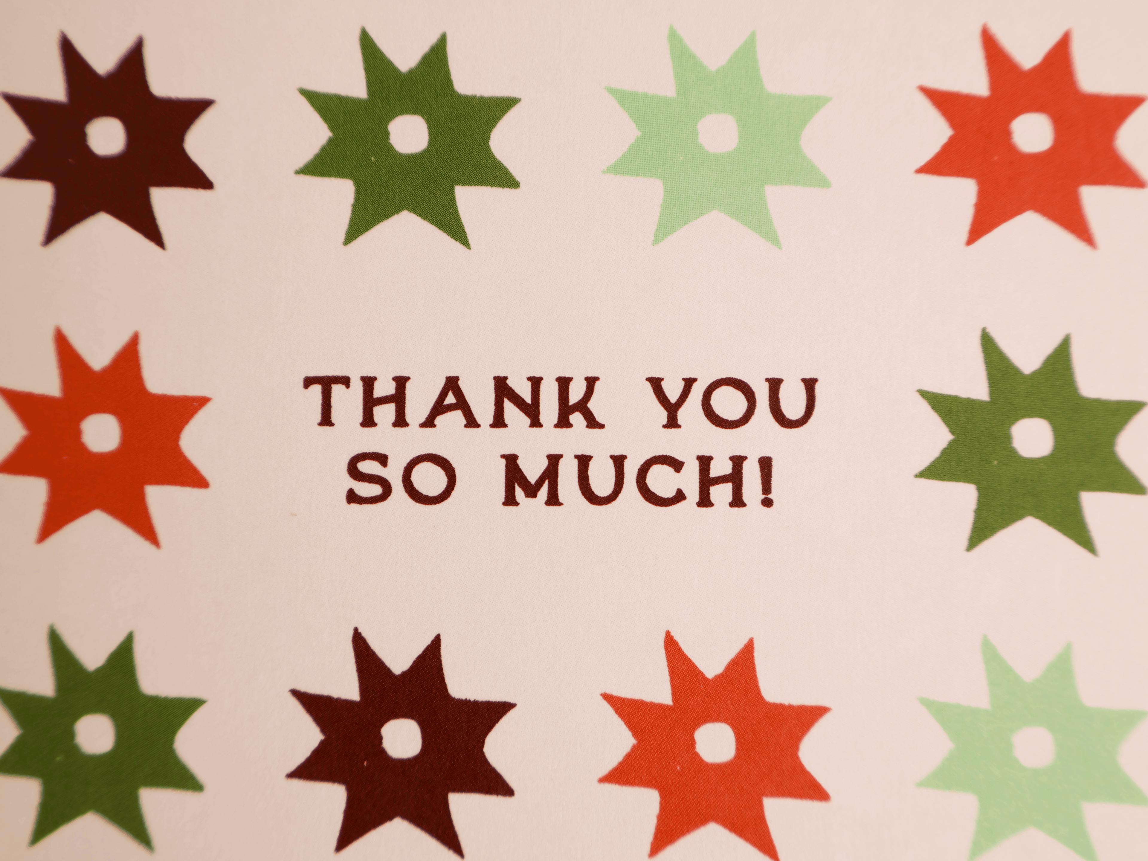 A close-up of a printed card or paper with the words “THANK YOU SO MUCH!” in dark red centered on a light background, surrounded by repeated starburst shapes in dark red, green, and pale mint.