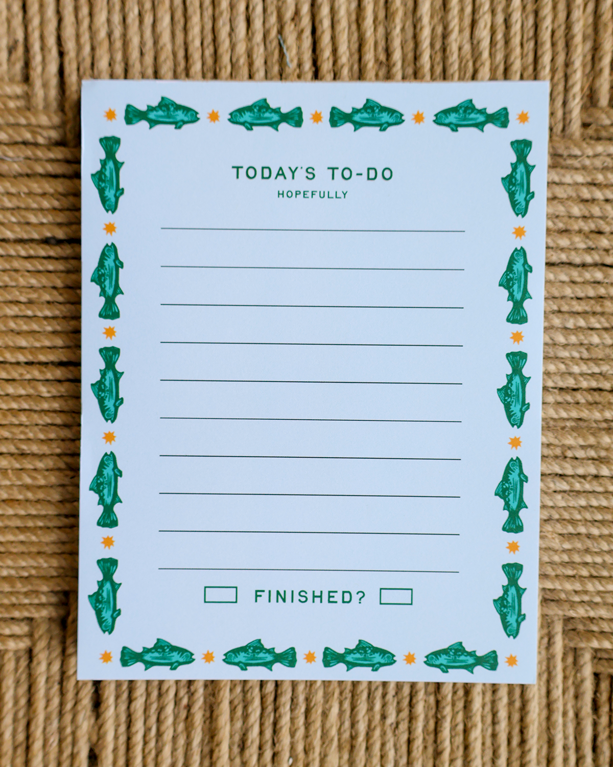A white notepad with a green fish border and the text 'TODAY'S TO-DO HOPEFULLY' and 'FINISHED?' printed on it, laid flat on a woven tan surface.