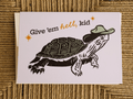 A square greeting card featuring a black and white illustrated turtle wearing a green hat, with the text 'Give 'em hell, kid' in black and orange lettering, placed on a woven tan rope surface.
