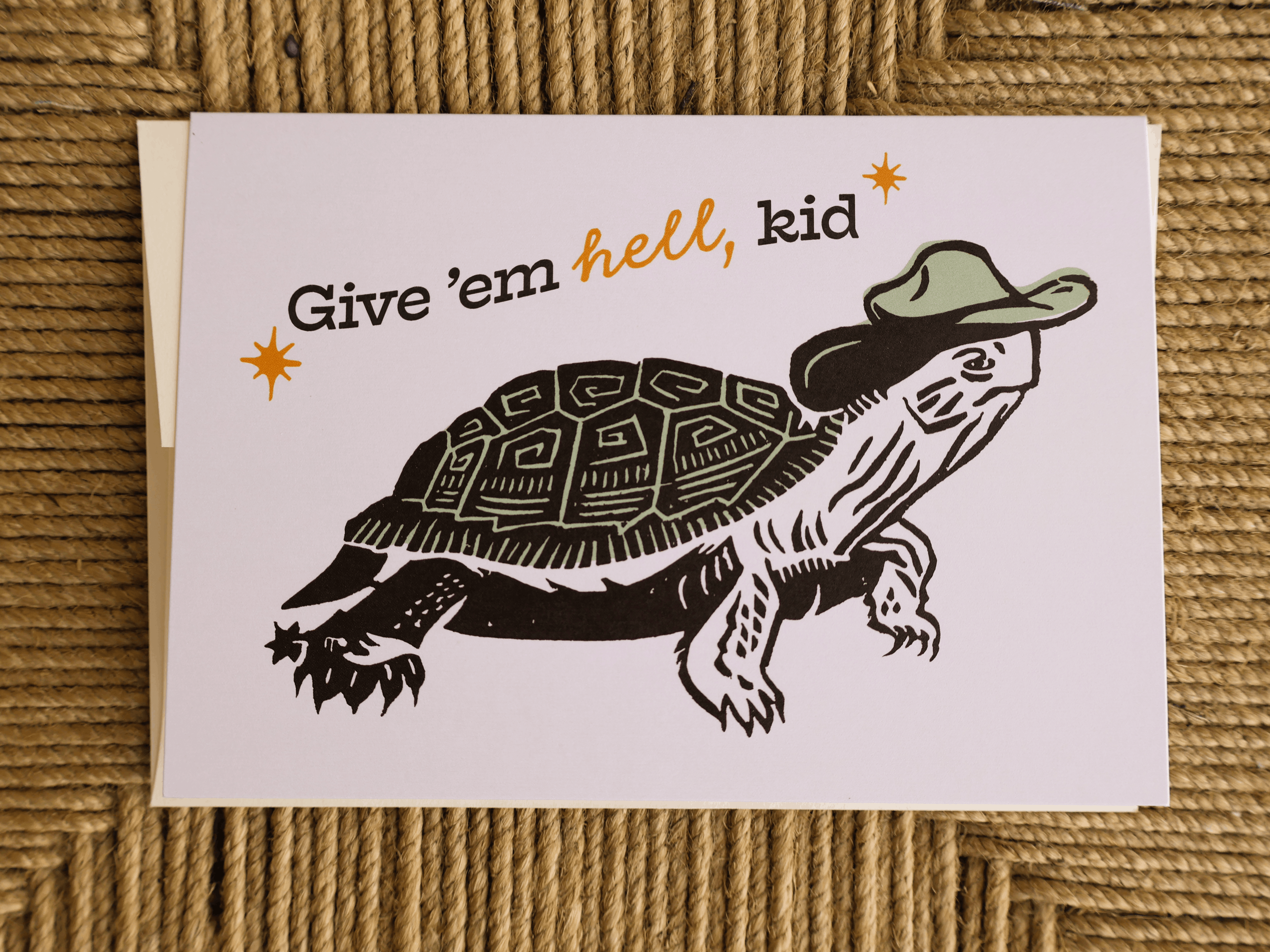 A square greeting card featuring a black and white illustrated turtle wearing a green hat, with the text 'Give 'em hell, kid' in black and orange lettering, placed on a woven tan rope surface.