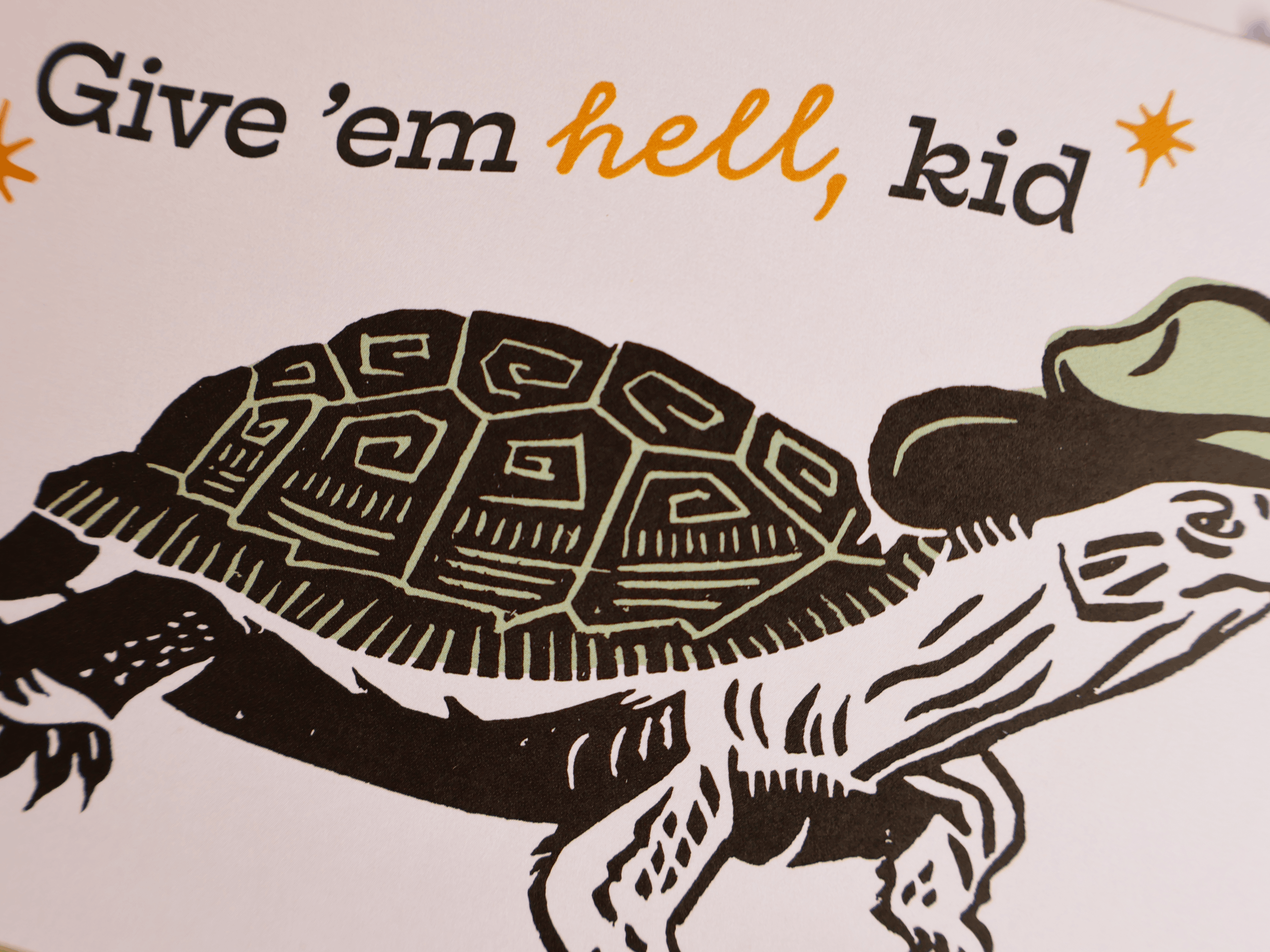 A close-up of a printed graphic with the text 'Give 'em hell, kid' in black and orange lettering above a black-and-white illustrated turtle wearing a light green hat on a white background.