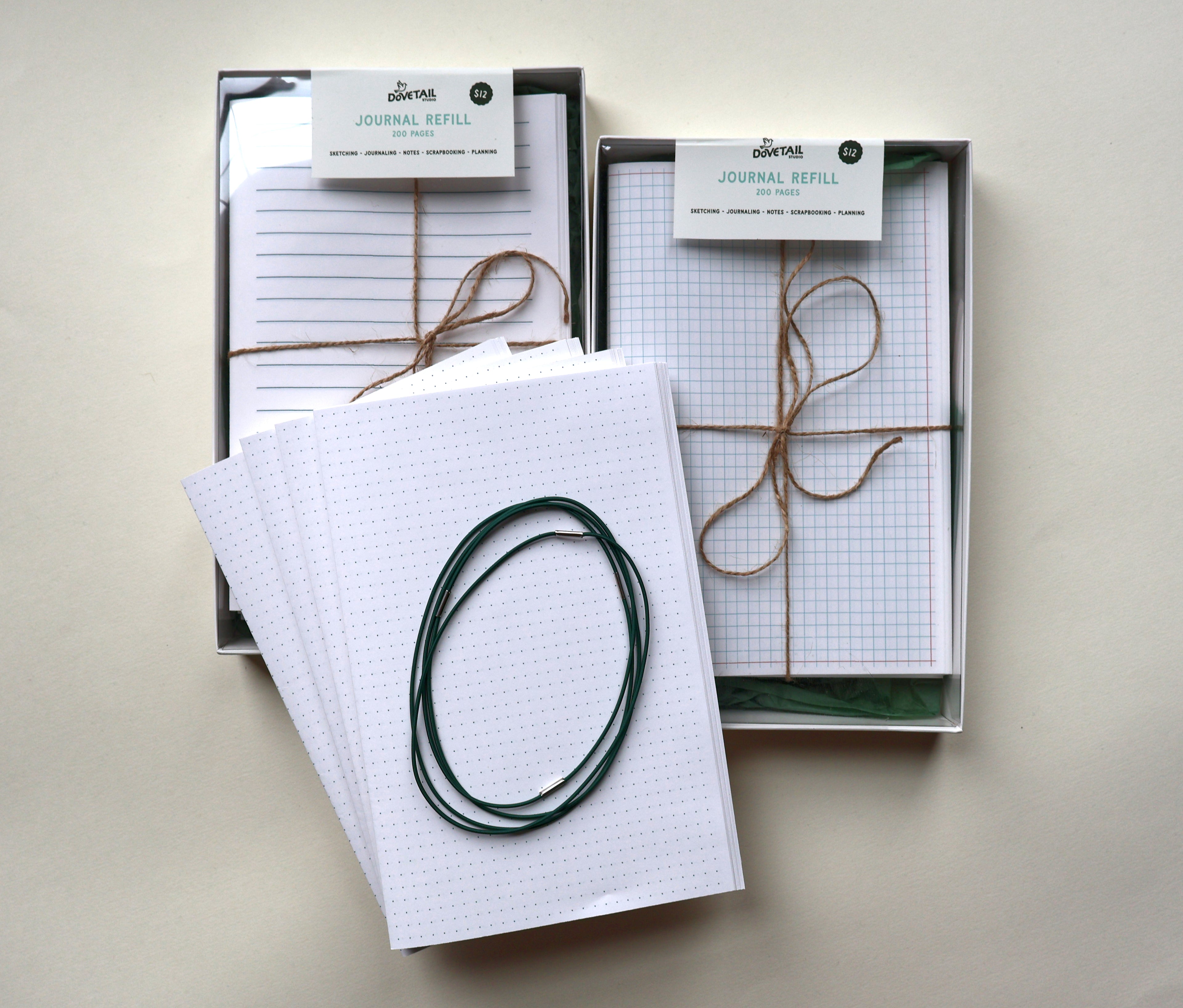 Two boxed paper journal refills tied with twine, shown on a light beige background. One box contains lined paper and the other contains graph paper, with a stack of white bullet paper and a dark green looped journal binding elastics placed in front.