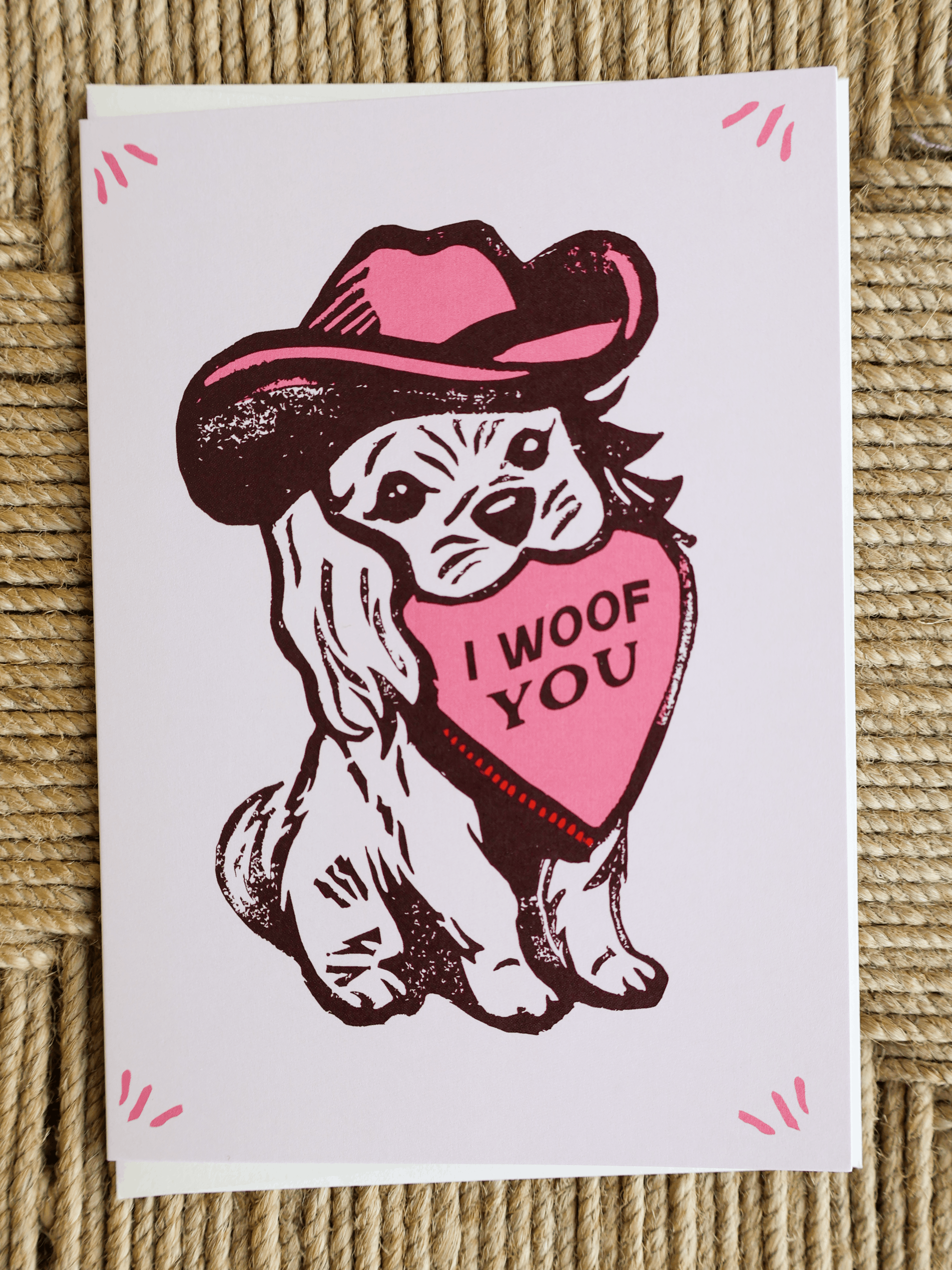A square greeting card featuring a cartoon dog wearing a pink cowboy hat and holding a pink heart that says 'I WOOF YOU', photographed on a woven beige surface.