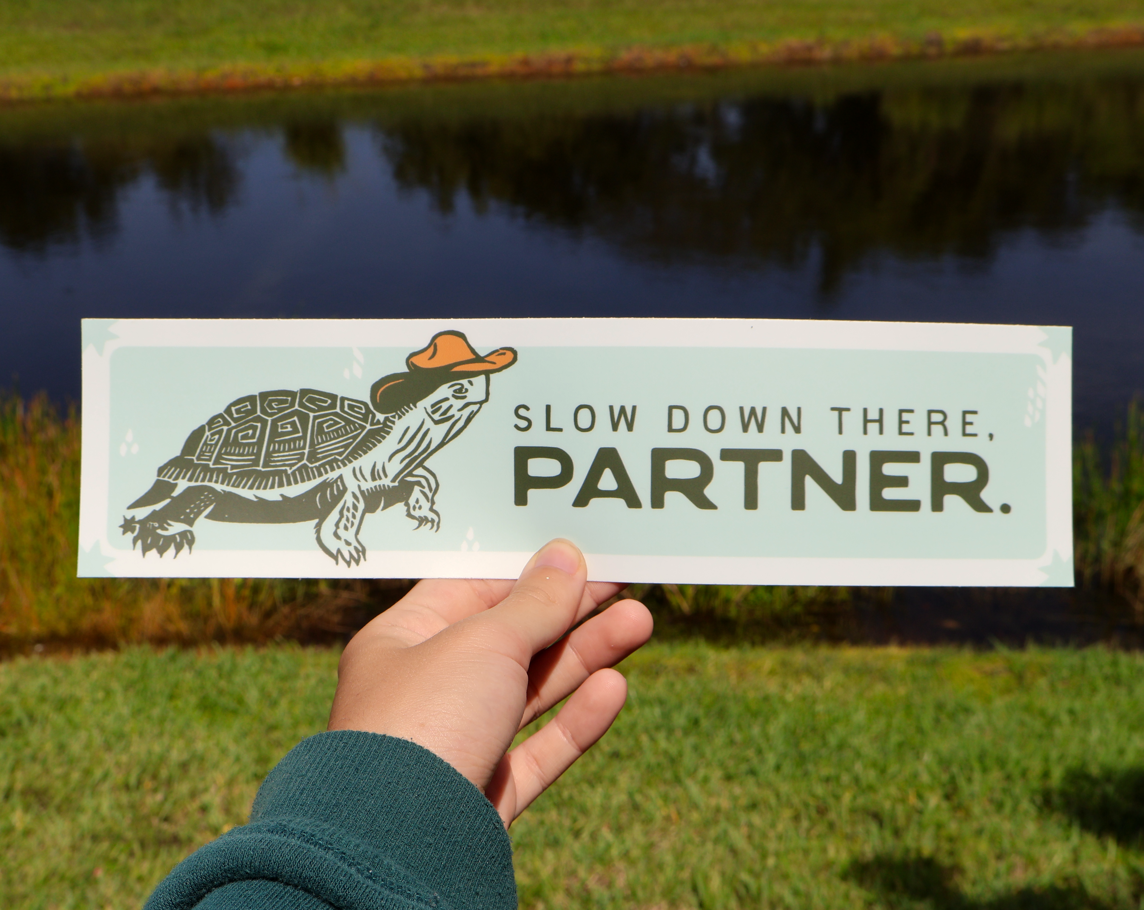 vinyl bumper sticker with a turtle wearing a cowboy hat and text 'Slow down there, partner' with lake and trees in the background