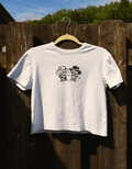 A white short-sleeve t-shirt hanging on a wooden hanger against a dark wooden fence outdoors. The shirt features a black graphic print of two cartoon fish wearing cowboy hats facing each other.