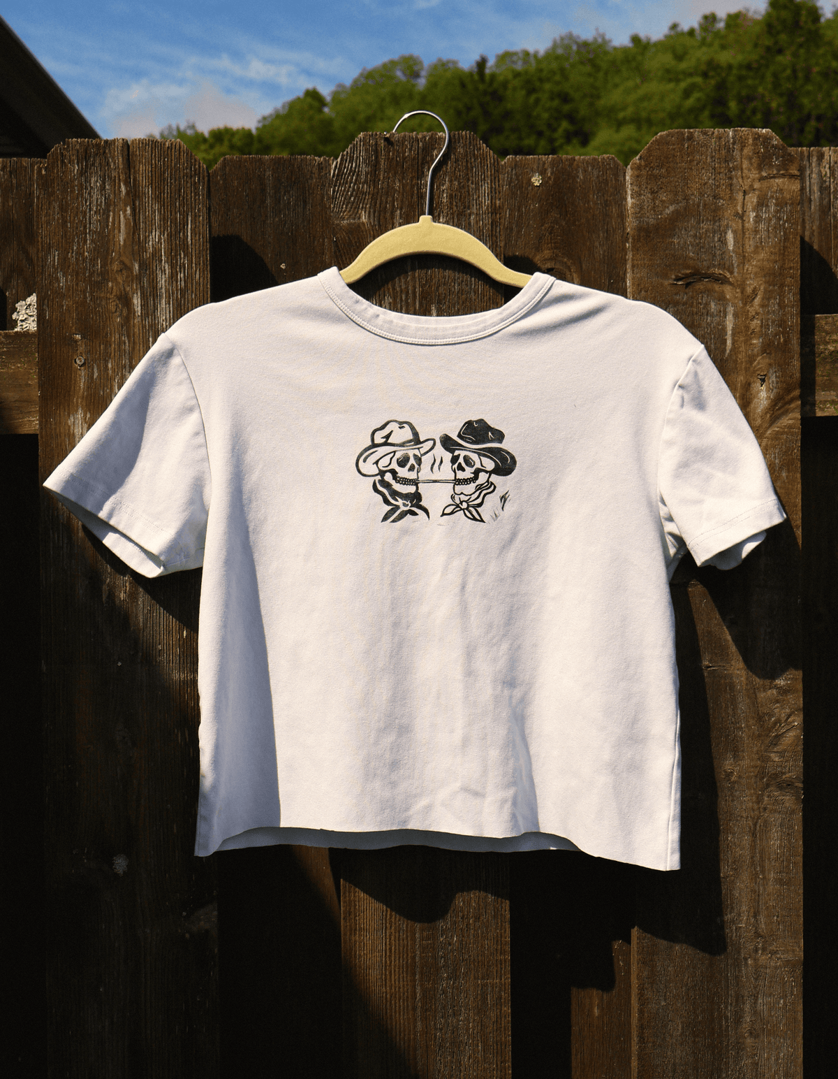 A white short-sleeve t-shirt hanging on a wooden hanger against a dark wooden fence outdoors. The shirt features a black graphic print of two cartoon fish wearing cowboy hats facing each other.