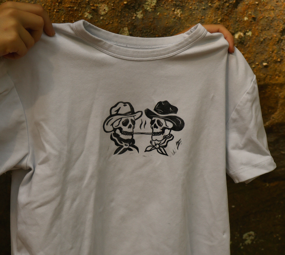 A white short-sleeve t-shirt with a black graphic of two cowboy-style skeletons holding a bow and arrow, held up by hands against a textured brown background.