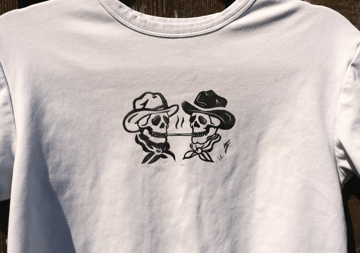 A white short-sleeve t-shirt featuring a black graphic print of two cowboy skulls wearing hats and smoking a joint.