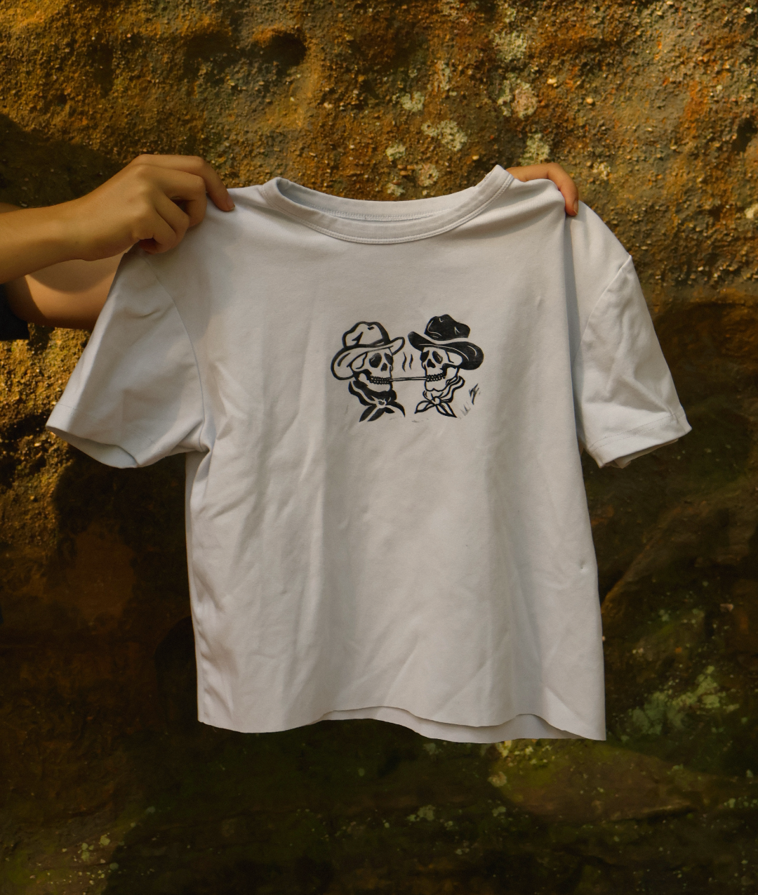A white short-sleeve t-shirt with a black graphic of two cartoon frogs wearing cowboy hats, held up by two hands against a textured, mossy brown rock wall.