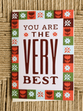 A square greeting card with the phrase 'YOU ARE THE VERY BEST' in dark brown lettering on a white background, surrounded by a colorful retro border of red, green, orange, and light blue floral and geometric motifs, placed on a woven beige surface.
