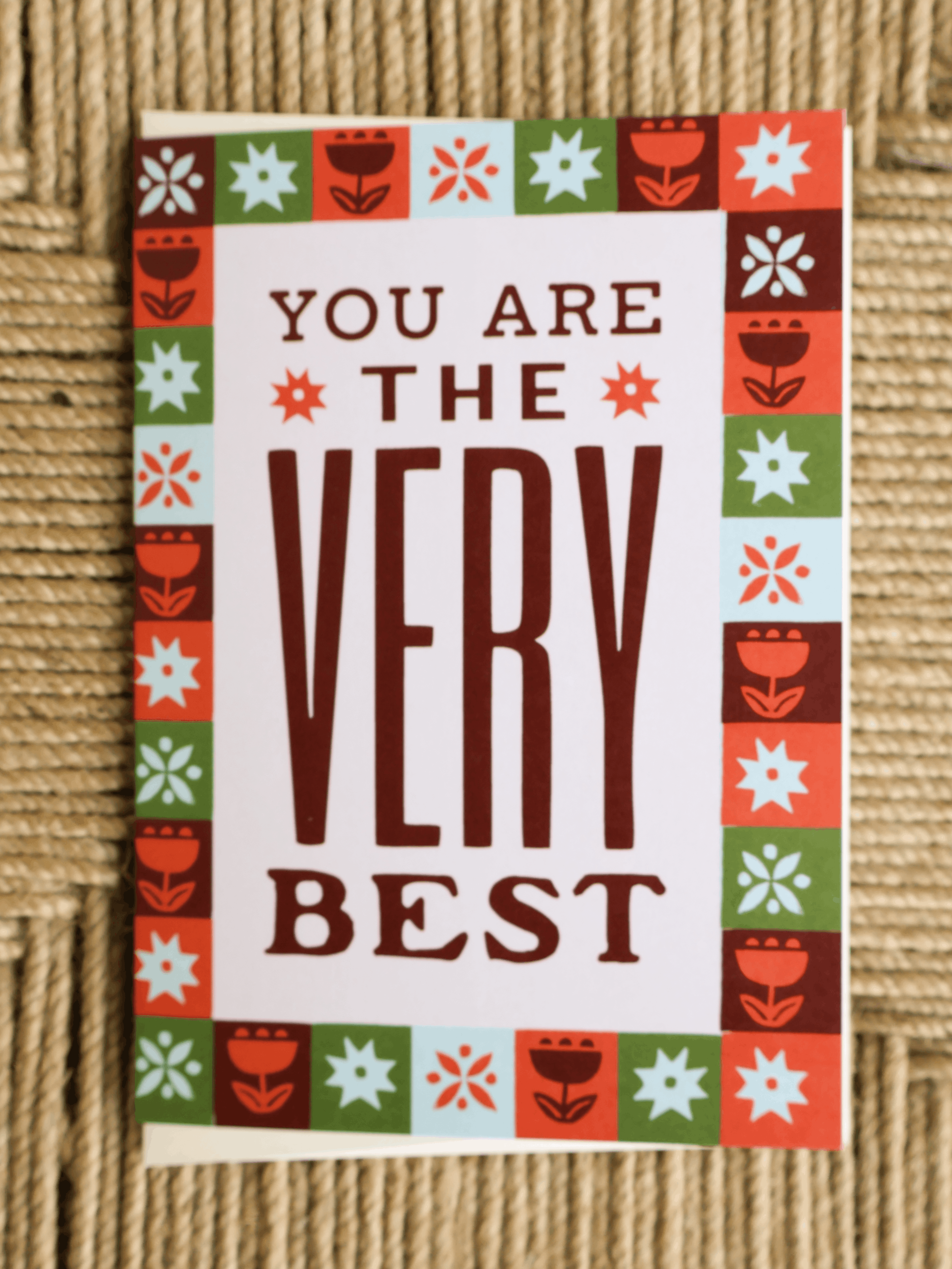 A square greeting card with the phrase 'YOU ARE THE VERY BEST' in dark brown lettering on a white background, surrounded by a colorful retro border of red, green, orange, and light blue floral and geometric motifs, placed on a woven beige surface.