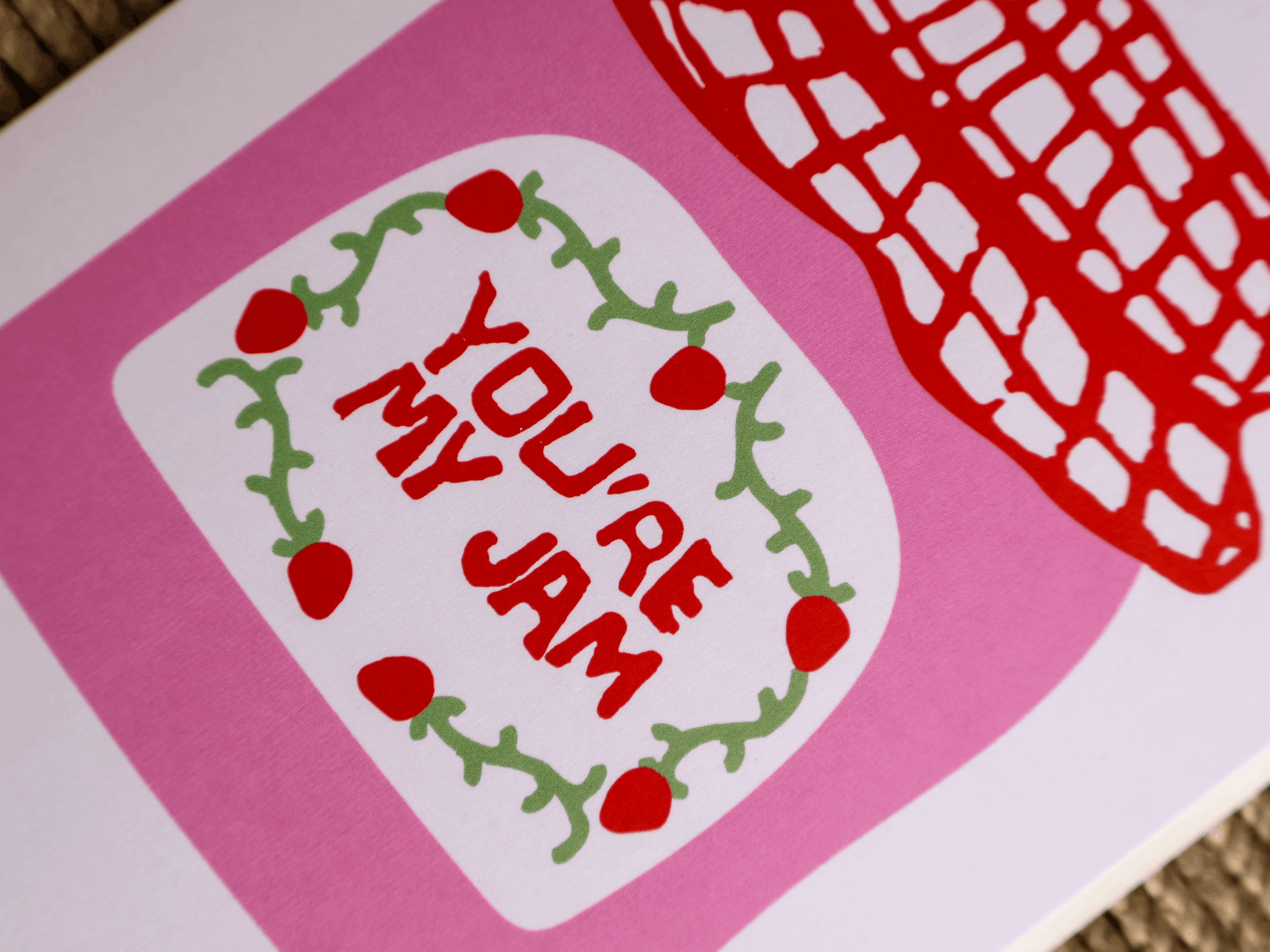 A close-up of a printed card or art piece with a pink background, a white label area reading “YOU’RE MY JAM” in red lettering, a green vine wreath with red berries, and part of a red patterned shape at the top right.