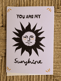 A white greeting card featuring a black sun illustration with a face and the text 'YOU ARE MY Sunshine' on a woven straw background.