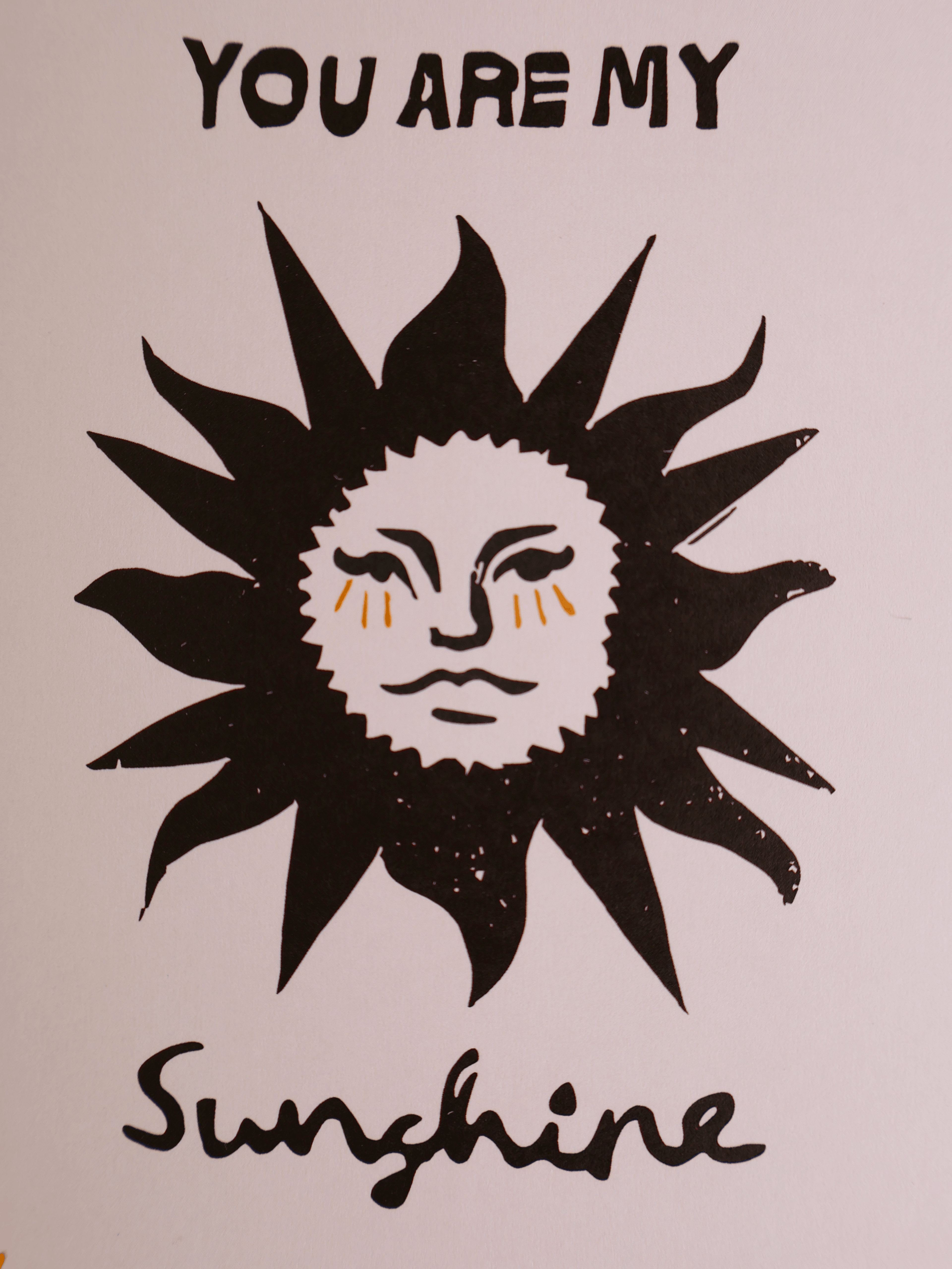 A square art print featuring a stylized black sun with a white face and small orange tear details, with the text 'YOU ARE MY Sunshine' in black on a light pink background.